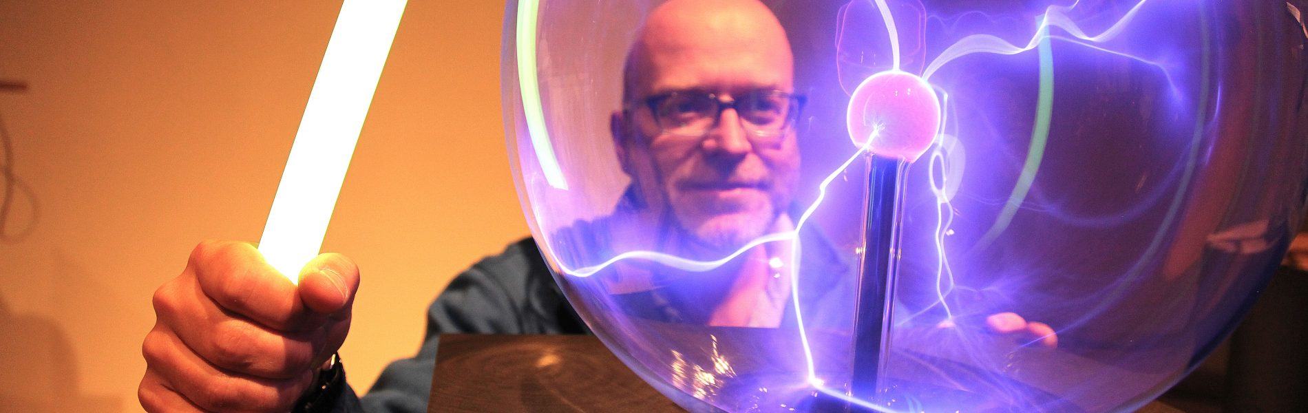 Das Bild zeigt einen Mann vor einer Plasmakugel. Die gläserne Kugel ist von der Mitte zum Rand mit kleinen, lilanen Blitzen durchzogen.