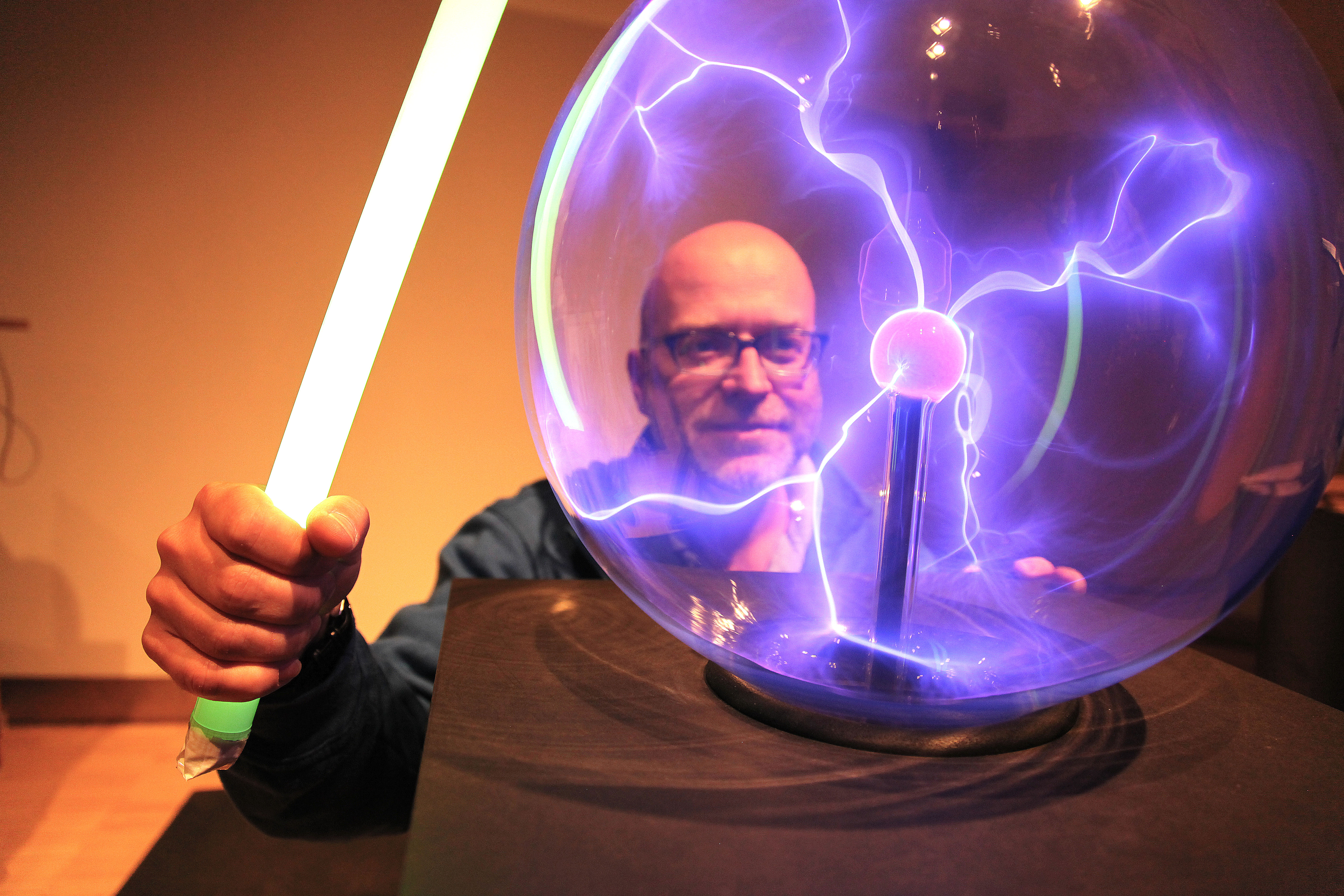 Das Bild zeigt einen Mann vor einer Plasmakugel. Die gläserne Kugel ist von der Mitte zum Rand mit kleinen, lilanen Blitzen durchzogen.